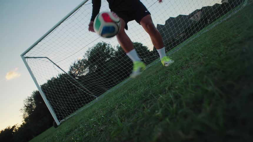 Low angle slow motion shot of a soccer player's legs as he skillfully juggles a ball on a grassy field in front of a goal at dusk
