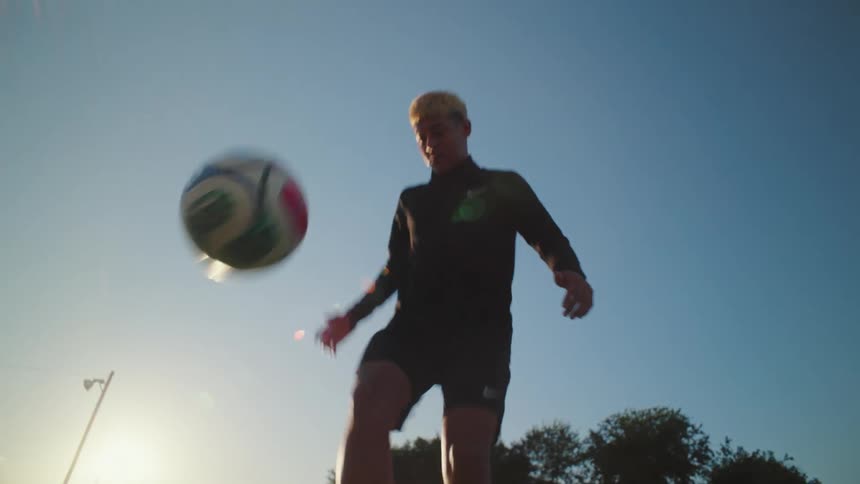 A skilled young soccer player juggles a soccer ball in slow motion from a low-angle perspective against the setting sun