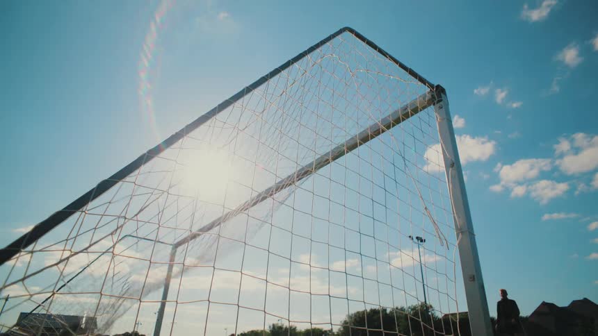 From a low angle, a soccer ball hits the back of the net in slow motion as the sun shines brightly