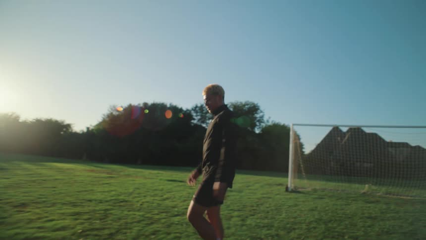 In slow motion, a young man with blonde hair practices juggling a soccer ball on a green field at sunrise