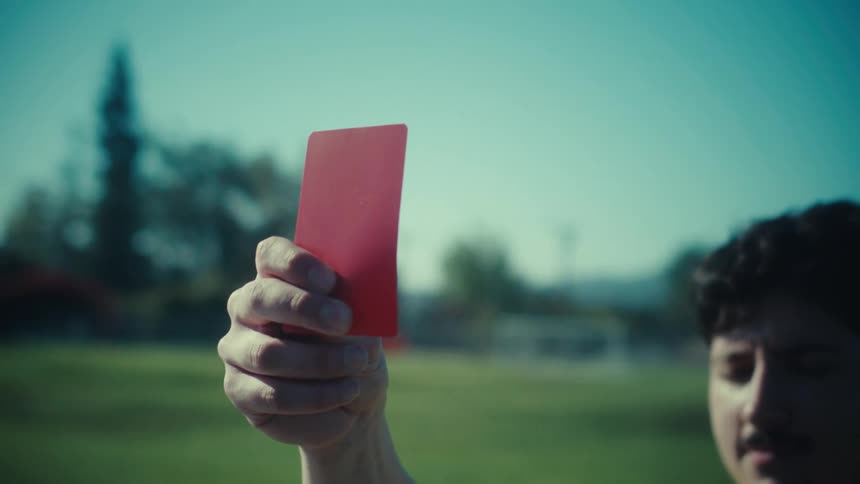 A serious soccer referee holds up a red card in slow motion on a sunny day at the field