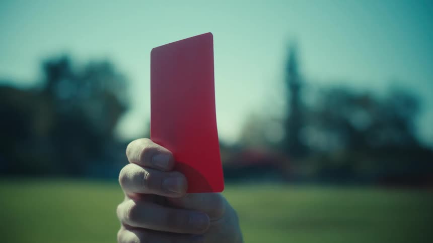 A referee's hand slowly comes into focus while holding up a red penalty card on a sports field