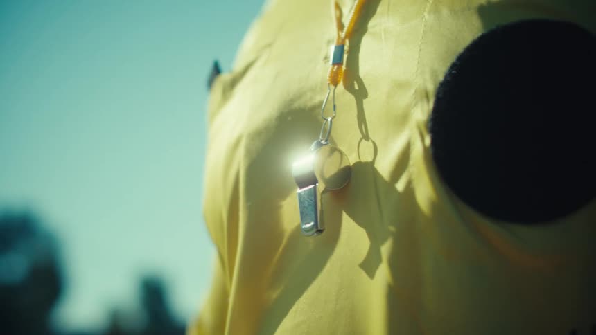 Dramatic slow-motion close-up of a referee's whistle hanging on a yellow jersey as the sun glints off of it
