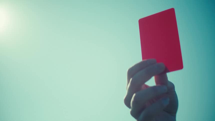 A football referee shows a red card, with the camera racking focus from the referee to the card against a sunny sky