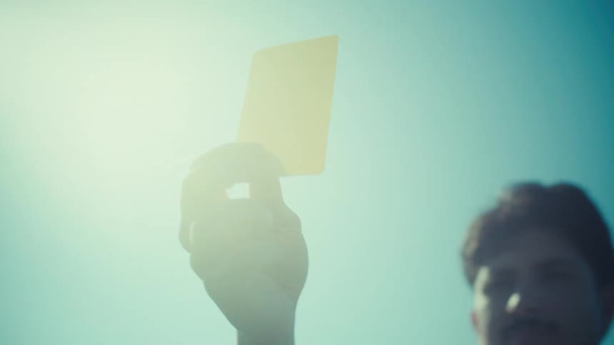 A referee's hand holds up a yellow penalty card against a bright, sunny sky in slow motion