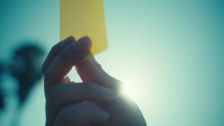 Dramatic slow-motion close-up of a referee's hand showing a yellow card against a sunlit sky