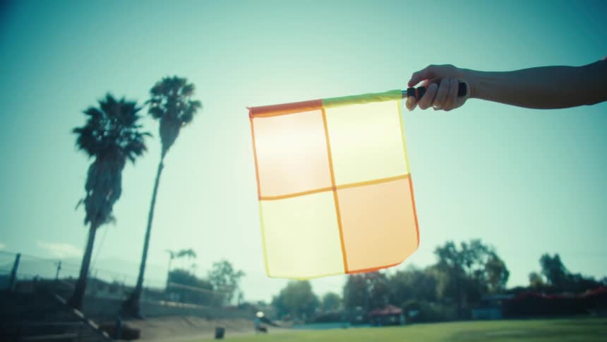 A referee's assistant holds up a yellow and orange checkered linesman flag on a sunny soccer field with palm trees