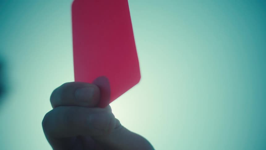 A referee's hand holds up a red card against the bright sun and a clear sky