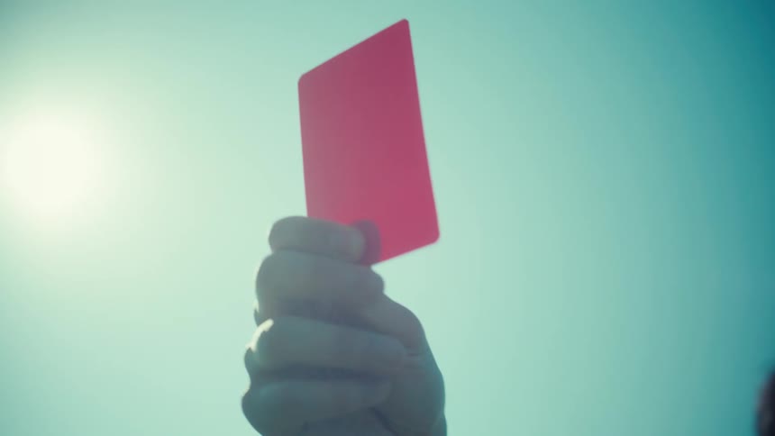 Dramatic low-angle view of a referee holding up a red card in slow motion against the sun