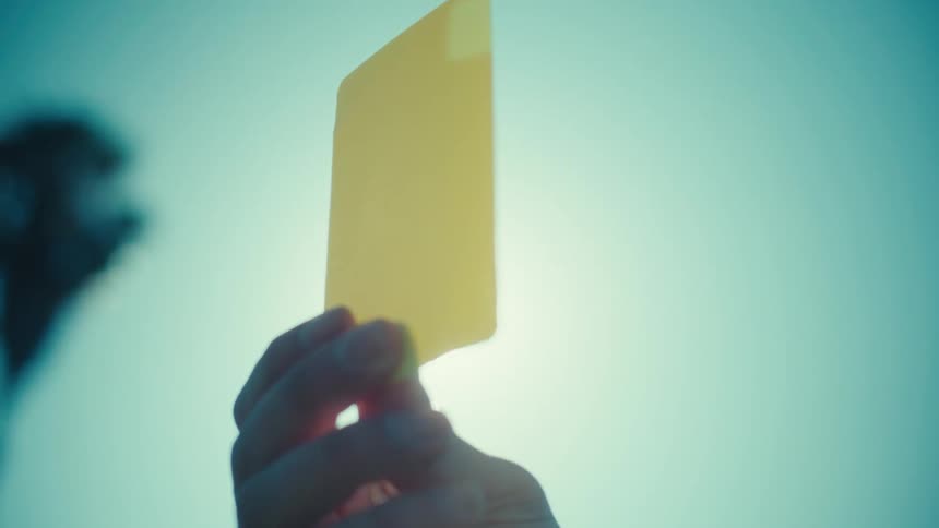 A referee's hand slowly raises a yellow penalty card against the bright, sunny sky