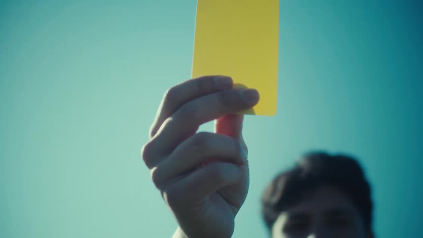 A serious referee holds up a yellow card as a warning against a bright, clear blue sky