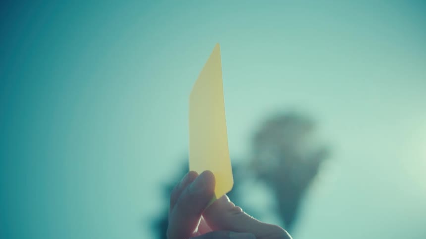 A referee's hand holds up a yellow penalty card against the bright sun in slow motion