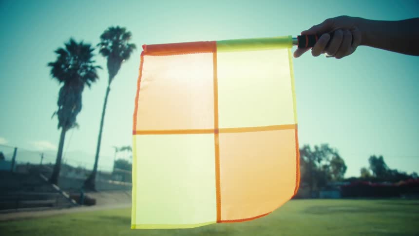 A linesman's checkered flag waves in slow motion on a sunny soccer field with palm trees in the background