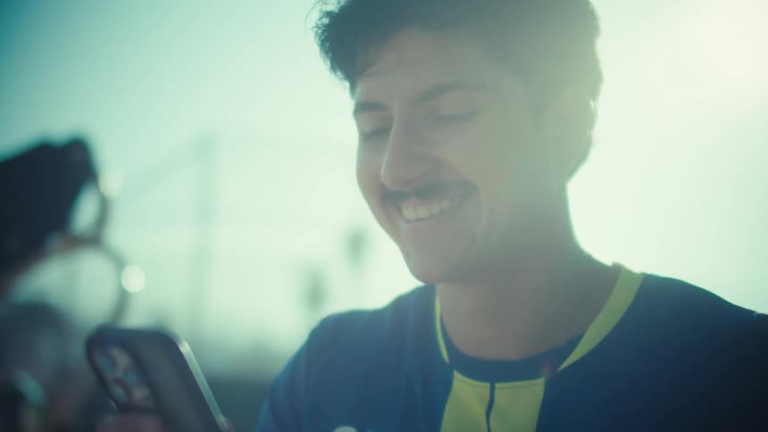 A young soccer player with a mustache smiles as he scrolls on his smartphone, backlit by the bright sun