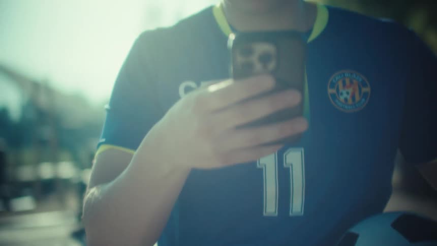 Close-up shot of a young soccer player scrolling on his phone while holding a football on a sunny day