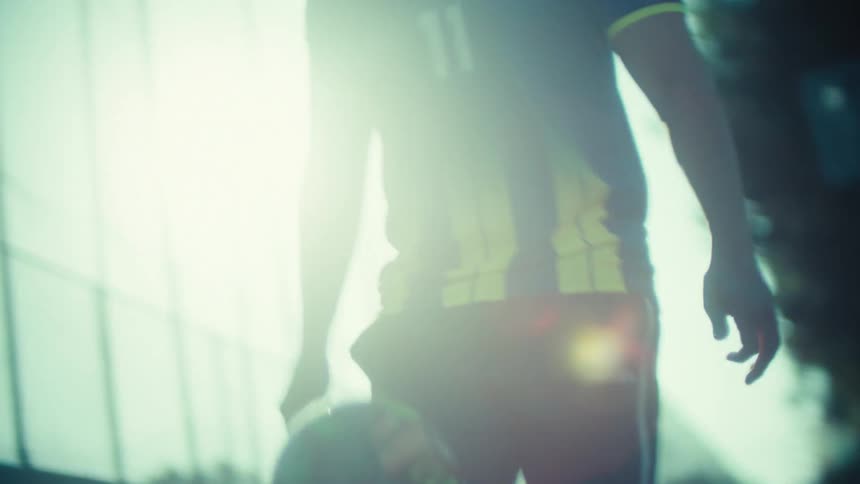 Slow-motion low-angle shot of a young soccer player skillfully juggling a ball while walking through a sunlit urban alleyway