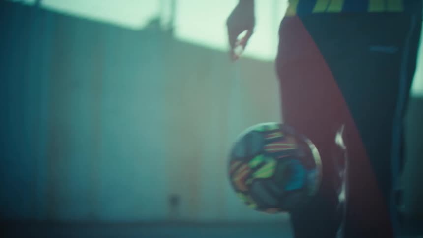 Stylized slow-motion close-up of a person skillfully juggling a soccer ball on a paved urban court with dramatic backlighting