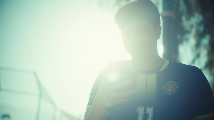 A young soccer player in a jersey smiles while looking at his phone with a bright sun flare behind him