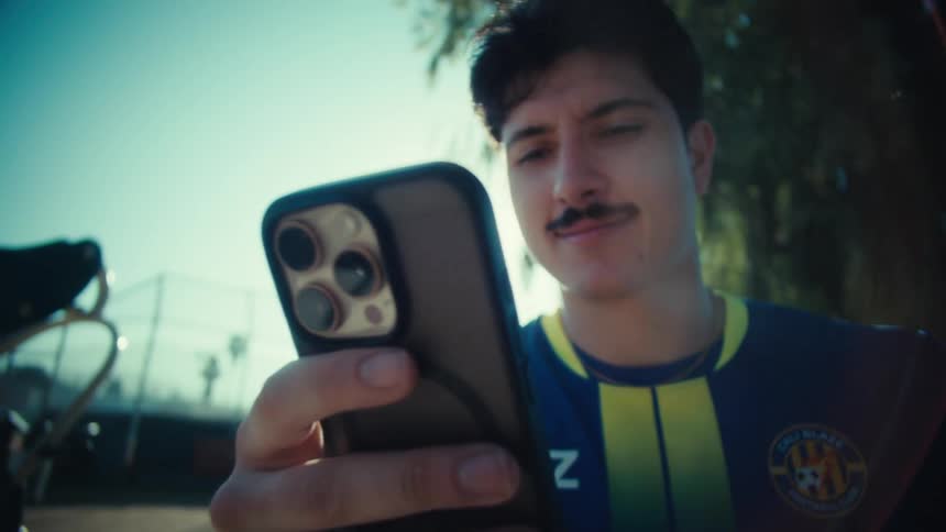 A young man in a soccer jersey smiles while using his smartphone outdoors on a bright sunny day