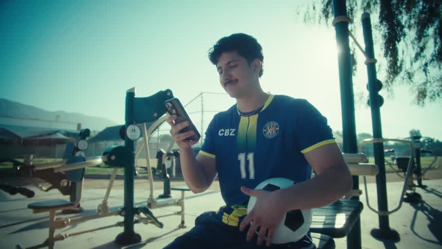 A young soccer player with a mustache smiles as he looks at his smartphone while resting at an outdoor gym in a sunny park