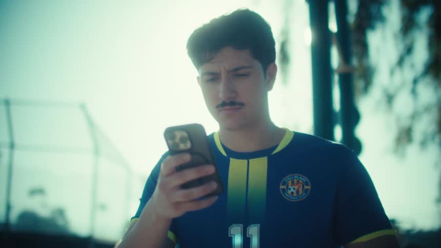 A young soccer player in uniform sits on a park workout bench, intently focused on his smartphone in the bright sunlight