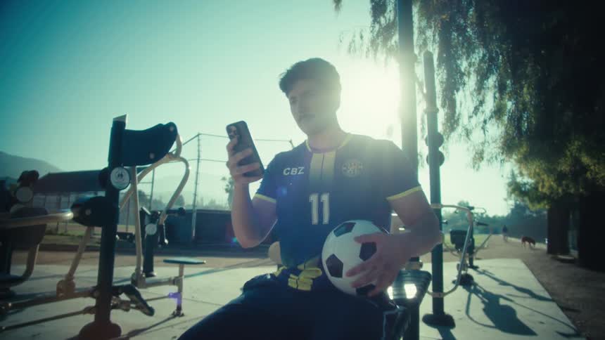 A young soccer player sits on outdoor gym equipment on a sunny day, scrolling on his smartphone as the camera slowly zooms in
