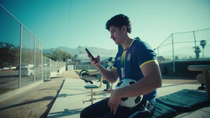 A young male soccer player in his team uniform scrolls on his smartphone while sitting on an exercise bench in a sunny park