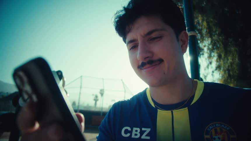 A smiling young man in a soccer jersey scrolls through his phone in slow motion at an outdoor park on a bright, sunny day