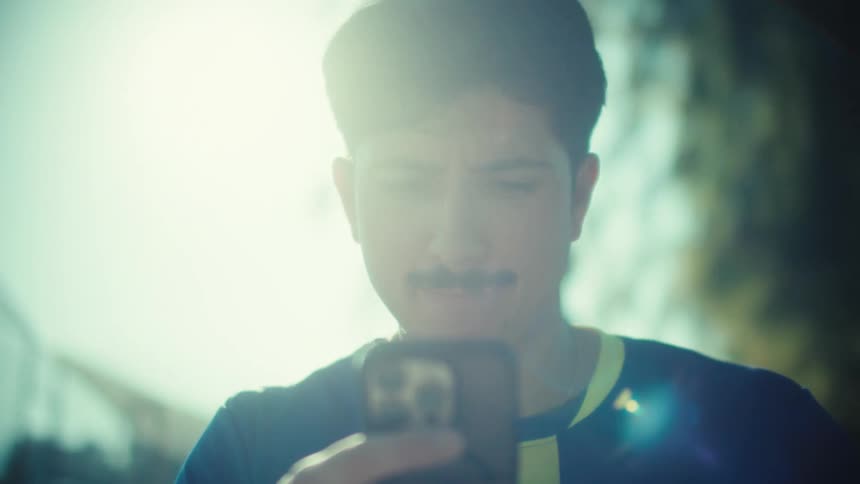 A young soccer player in his jersey sits on an exercise bench at a park, intently scrolling through his smartphone on a bright sunny day
