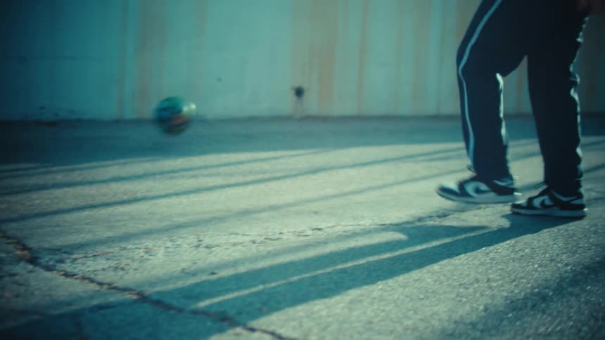 A person's feet skillfully juggle a soccer ball in slow motion on a cracked pavement court in a gritty urban setting