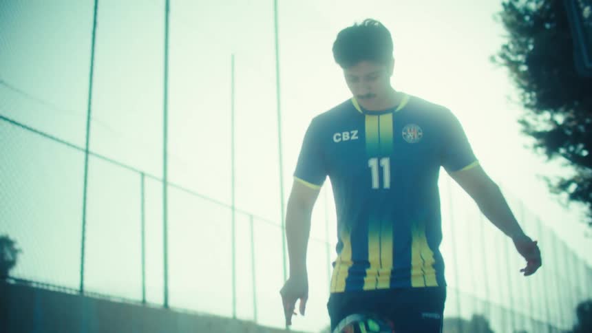A skilled young soccer player in a jersey practices juggling a ball in slow motion on a sunny, urban court