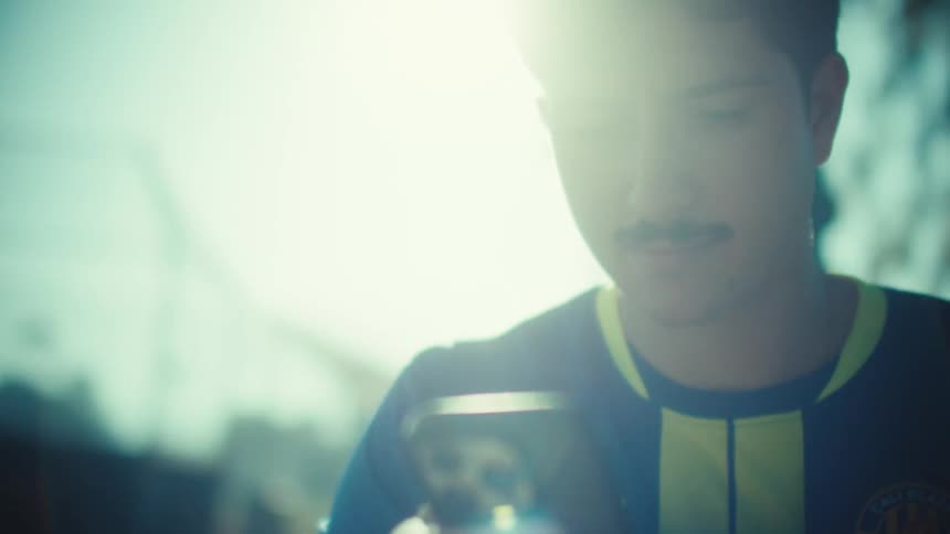 A thoughtful young man with a mustache in a sports jersey scrolls through his smartphone, backlit by a hazy sun flare
