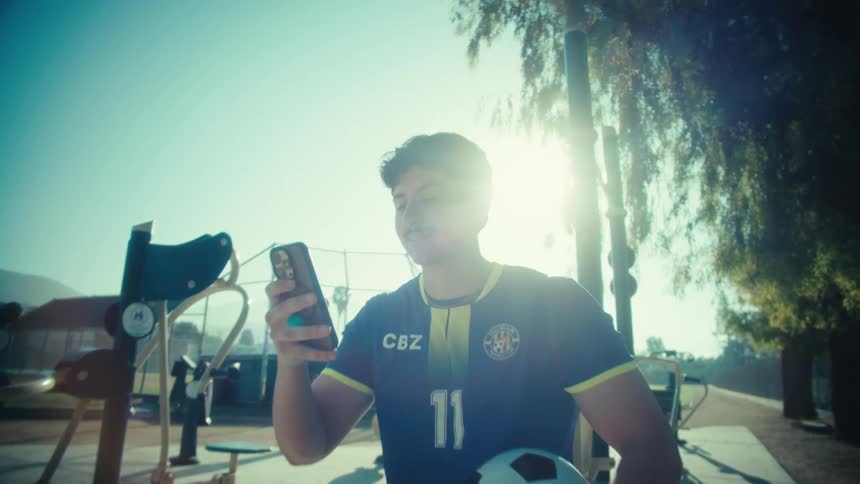 A young soccer player in his uniform smiles while scrolling on his smartphone at an outdoor gym in a sunny park