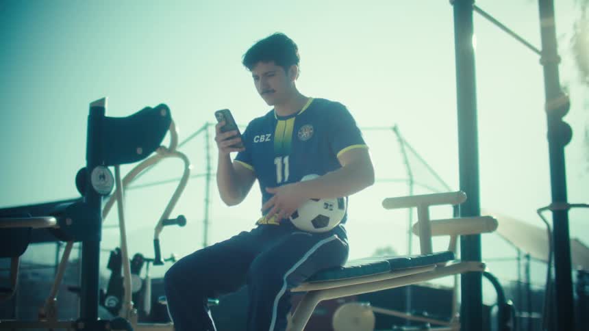 A young soccer player with a mustache takes a break at an outdoor gym to check his smartphone