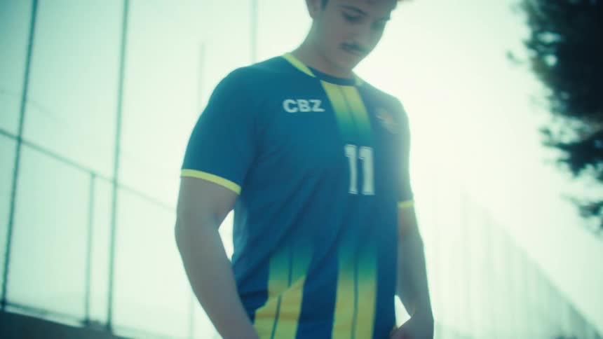 A skilled young soccer player in his jersey juggles a ball in slow motion on a sun-drenched urban court