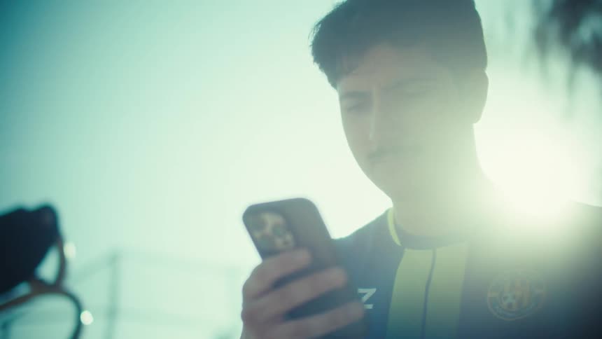 Slow-motion, low-angle close-up of a backlit soccer player with a mustache looking at his smartphone on a sunny day