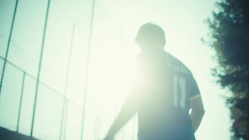 A skilled soccer player wearing a 'Captain' jersey practices juggling a ball in slow motion on an asphalt court against the bright sun