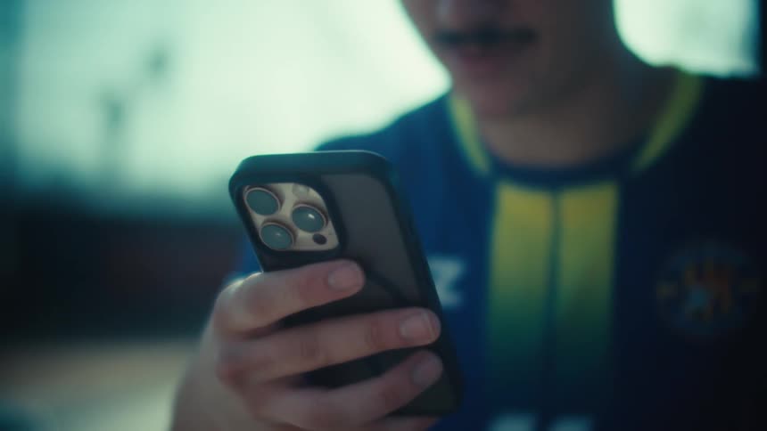 Close-up of a smiling young man in a soccer jersey reacting positively to something on his smartphone with bright sun flare in the background