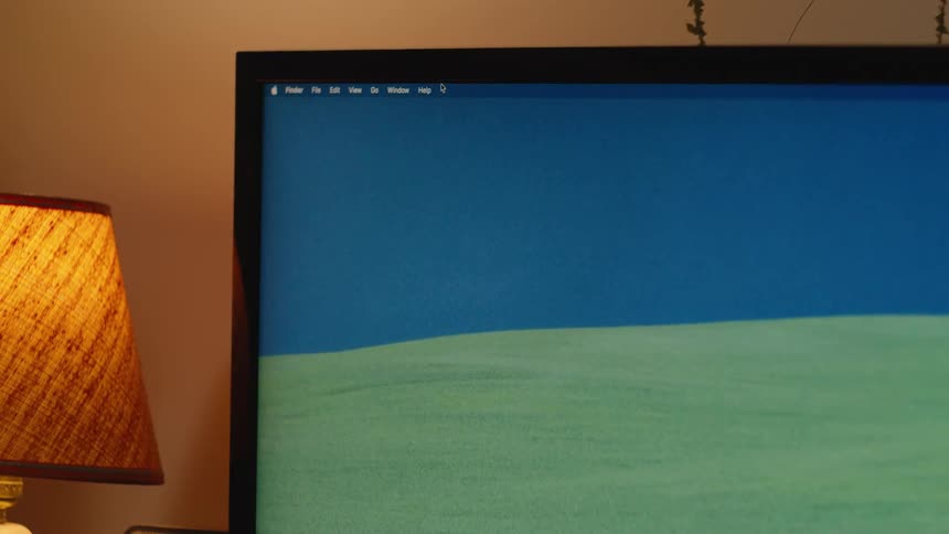 A close-up of a computer monitor displaying the macOS desktop with a classic landscape wallpaper next to a warm lamp