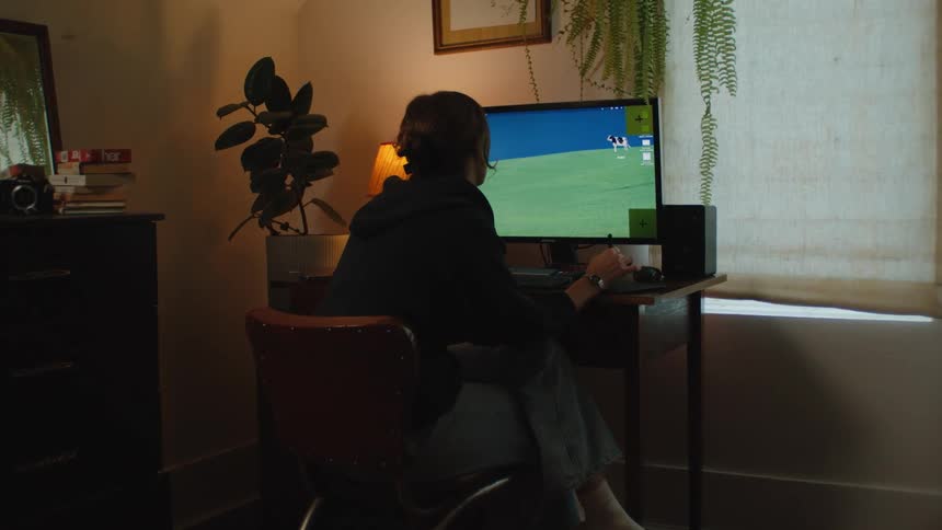 A female digital artist seen from behind works on a graphics tablet and computer in her cozy, dimly lit bedroom office