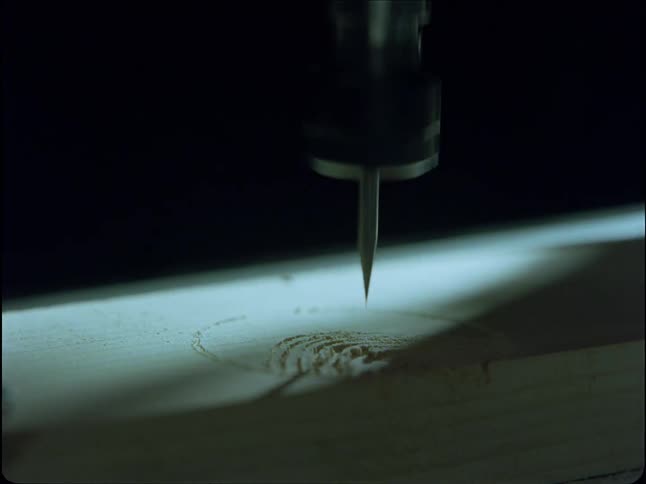 A CNC machine drill bit precisely carves a pattern into a block of wood under a single spotlight