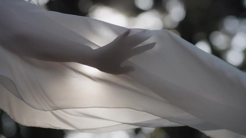 A hand's silhouette is seen through a sheer white fabric gracefully blowing in the wind in slow motion