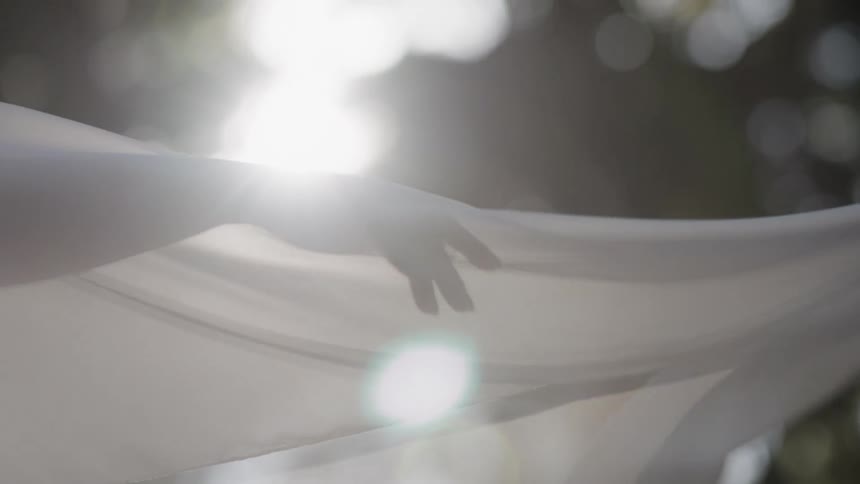 A silhouetted hand gently caresses a sheer white fabric blowing in the breeze as sunlight flares in slow motion