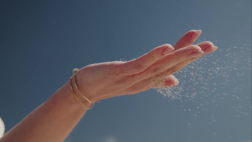 Slow motion close up of fine sand falling through a woman's fingers against a clear blue sky