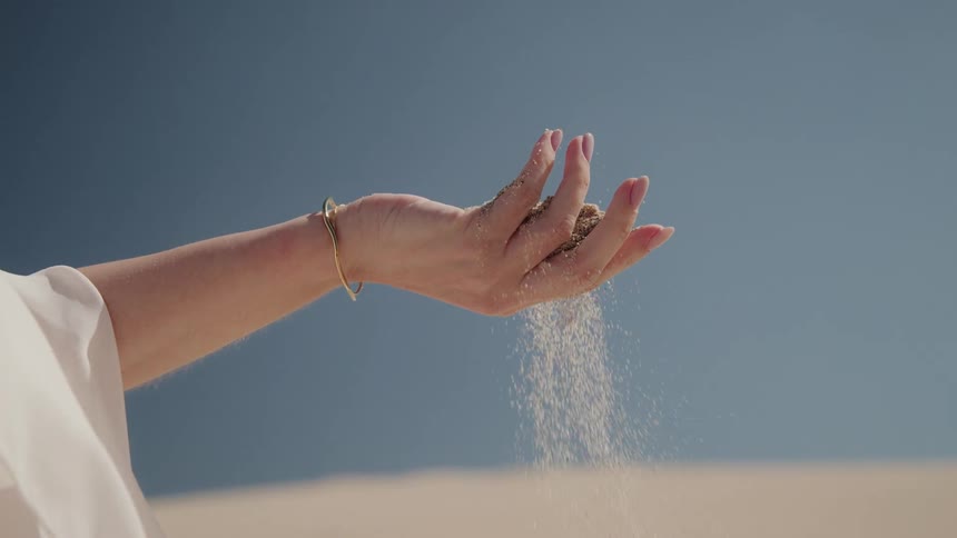 Close up of a woman's hand wearing a gold bracelet as sand slips through her fingers in slow motion against a desert backdrop