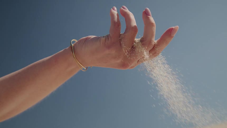 Slow motion close up of a woman's hand as sand elegantly slips through her fingers against a clear blue sky