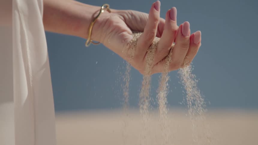 A closeup of a woman's manicured hands scooping up beach sand and letting it fall through her fingers in slow motion