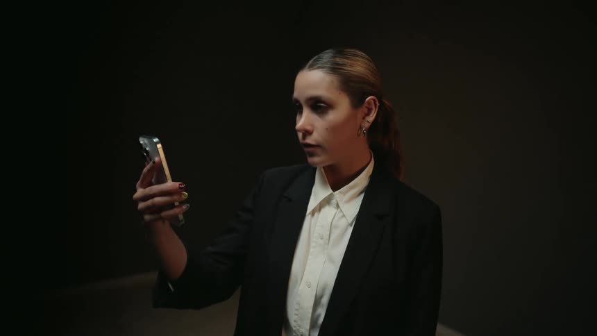A serious businesswoman in a dark suit raises her smartphone to look at the screen in a dimly lit room
