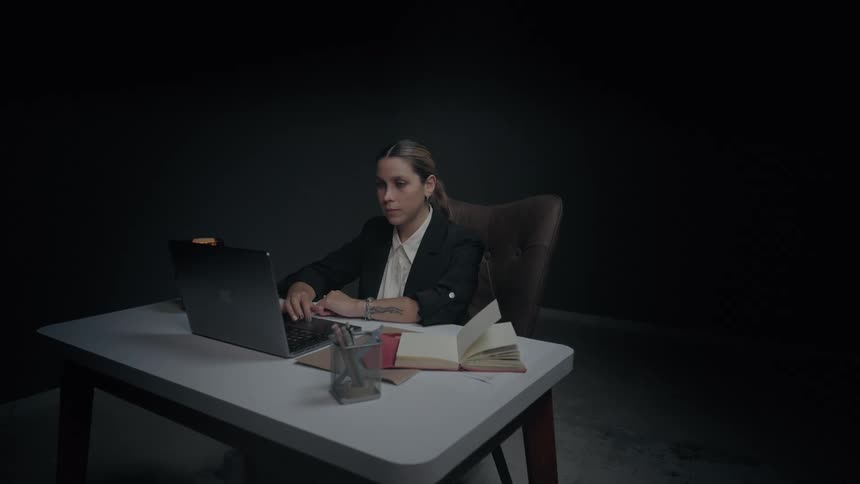 A professional woman in a suit works diligently on her computer late at night in a dark, minimalist office