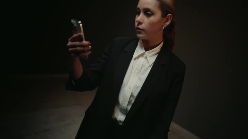 A woman in a black business suit lifts her smartphone and looks at the screen in a dark, moody room